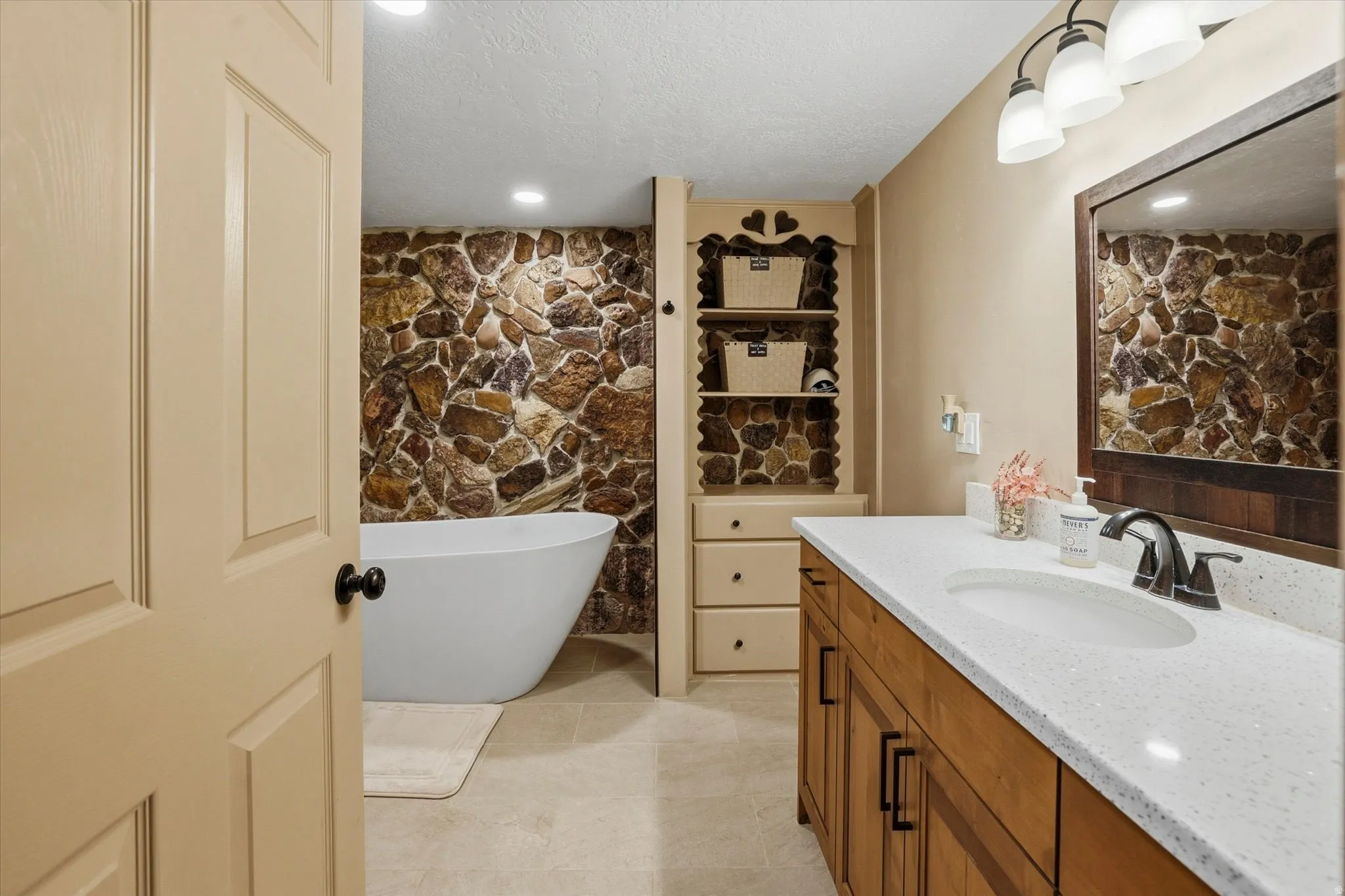 Freestanding soaking tub with a natural stone accent wall, complemented by a wood-finish vanity with a light-toned countertop