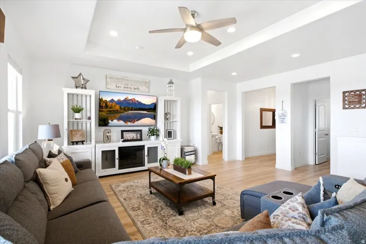 Spacious living area featuring light wood-finish flooring, a tray ceiling with recessed lighting, and a ceiling fan