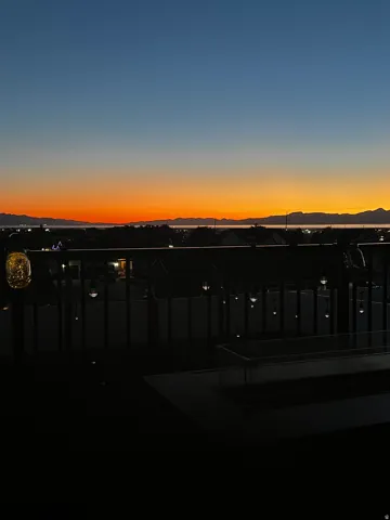 Outdoor balcony featuring a metal railing, string lights, and a tabletop fire pit, with a vibrant sunset backdrop over distant mountains