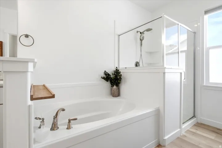 Built-in soaking tub with bronze-finish fixtures, enclosed shower with glass doors, wood-finish flooring, and a bright window