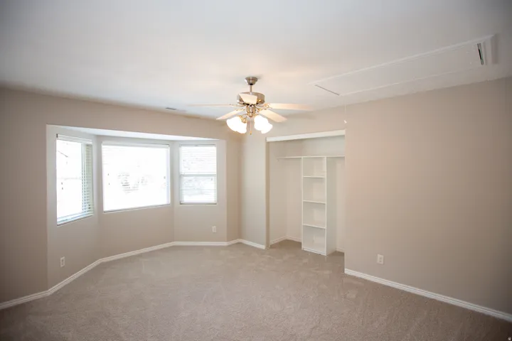 Neutral-toned room featuring a bay window, ceiling fan with light fixture, light-colored carpeting, and a built-in closet with shelving