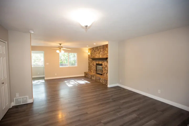 Spacious great room featuring wood-finish flooring, a stone-clad fireplace with a dark wood mantel, and multiple windows providing natural light