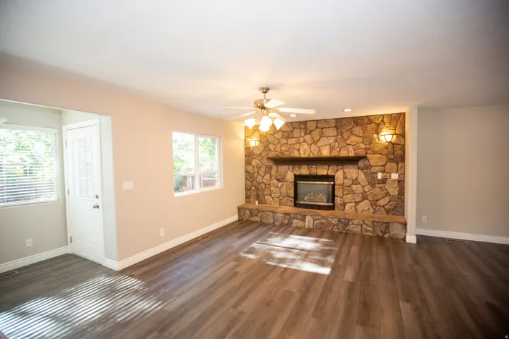 Spacious great room area featuring a prominent stone fireplace, wood-finish flooring, two large windows, a ceiling fan with integrated lighting, and sconce lighting