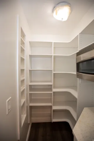 Walk-in pantry featuring extensive white shelving, wood-finish flooring, a built-in microwave, and a light-toned countertop