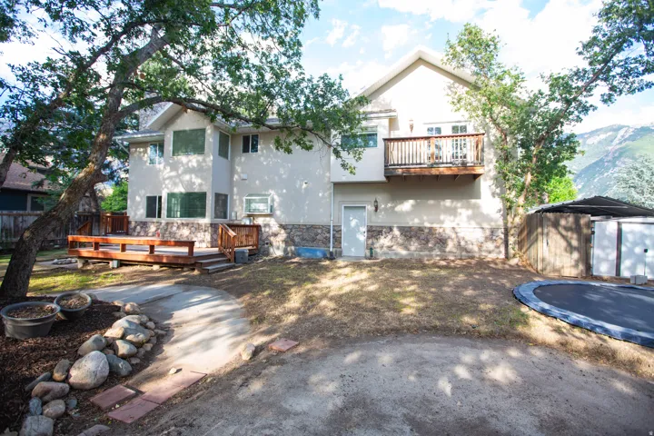 Rear exterior featuring a stucco and stone-finish facade, an upper-level balcony with wood railing, a ground-level wood deck, mature trees, and a mountain backdrop