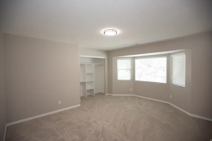 Bedroom featuring a bay window with blinds, neutral carpeting, light neutral wall paint, white baseboards, and a built-in closet system with shelving