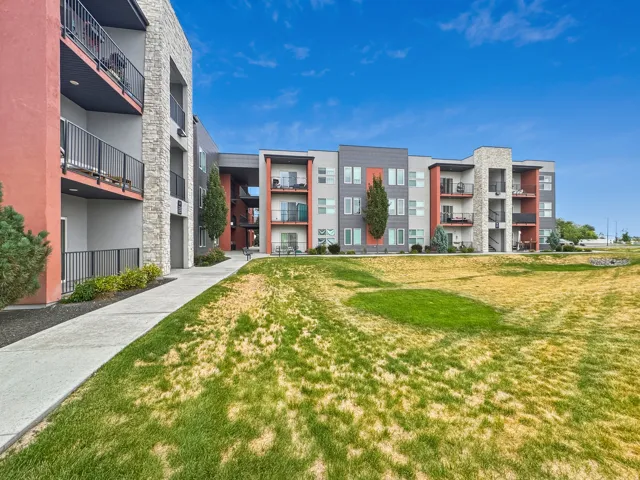 Contemporary multi-story building featuring stone and panel siding, private balconies with metal railings, and a paved pathway alongside a grassy area