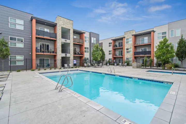Outdoor swimming pool with concrete decking, surrounded by multi-story buildings featuring balconies and a mix of exterior siding and stone accents