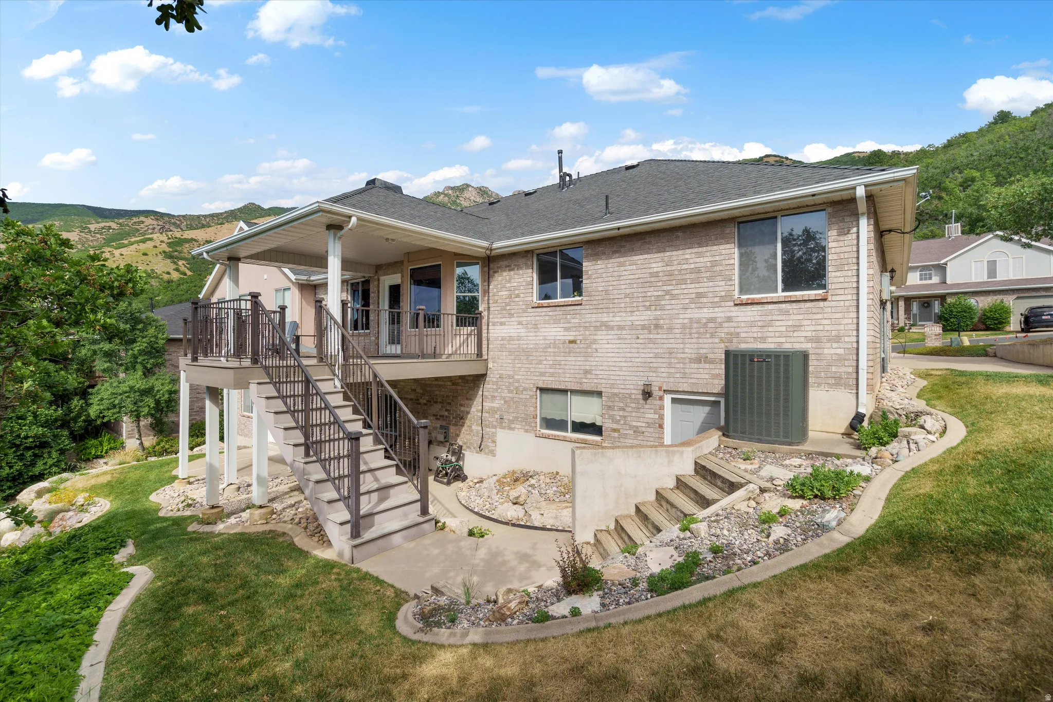Rear exterior featuring a brick facade, covered deck with railings, exterior staircase, and mature landscaping