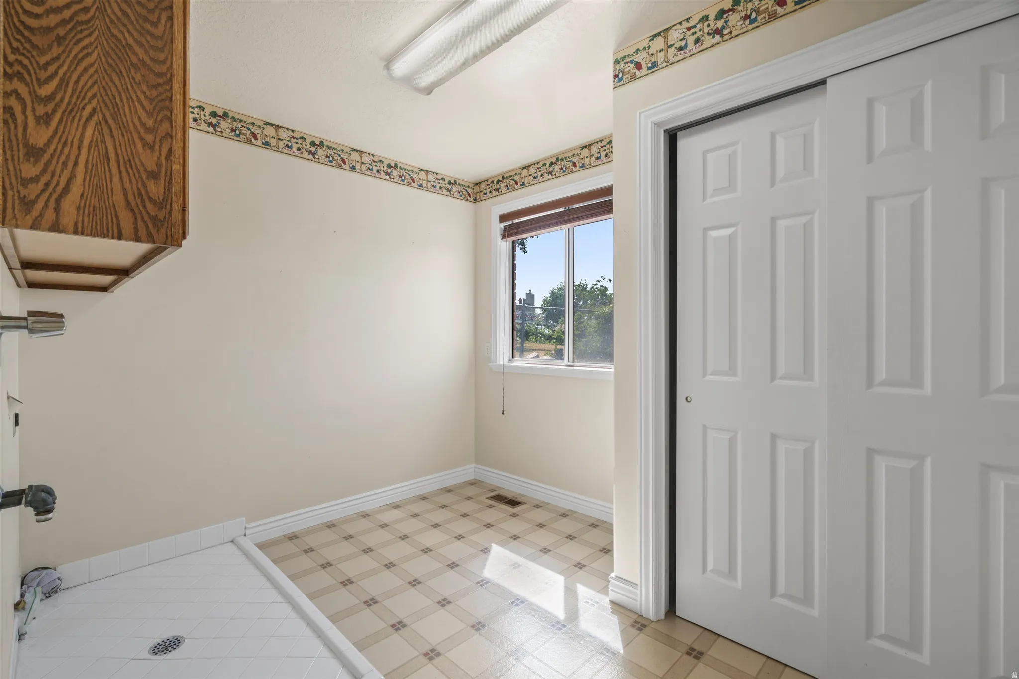 Utility room featuring a window with blinds, overhead fluorescent lighting, wood-finish cabinetry, a utility sink, and patterned floor tiling