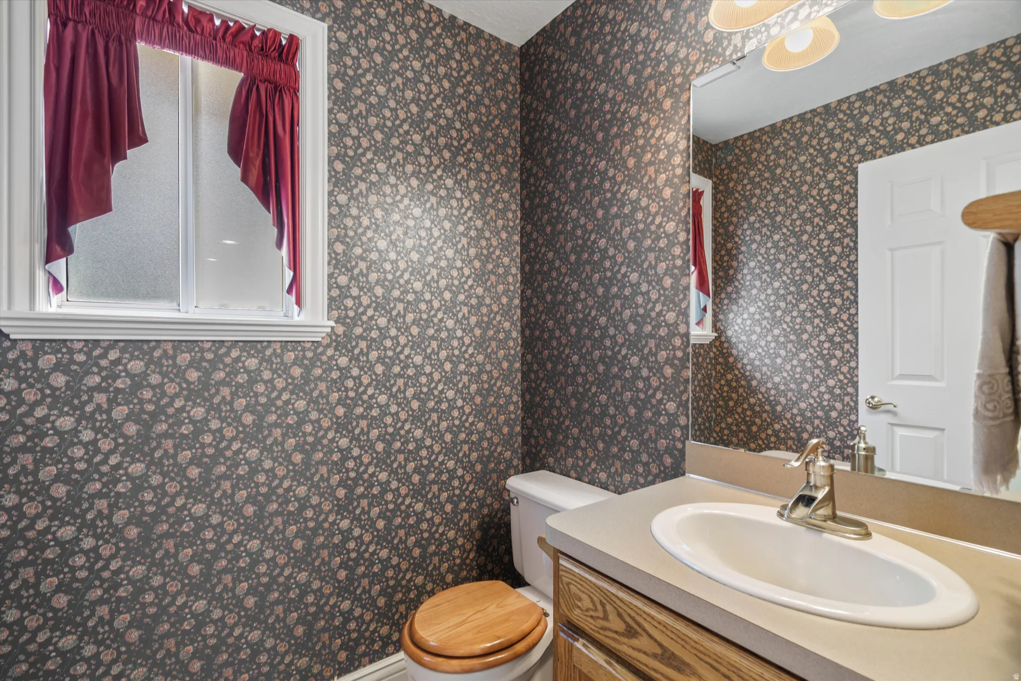 Powder room featuring a single-pane window, patterned wallpaper, a built-in vanity with an oval sink, a wood-finish cabinet, and a framed mirror