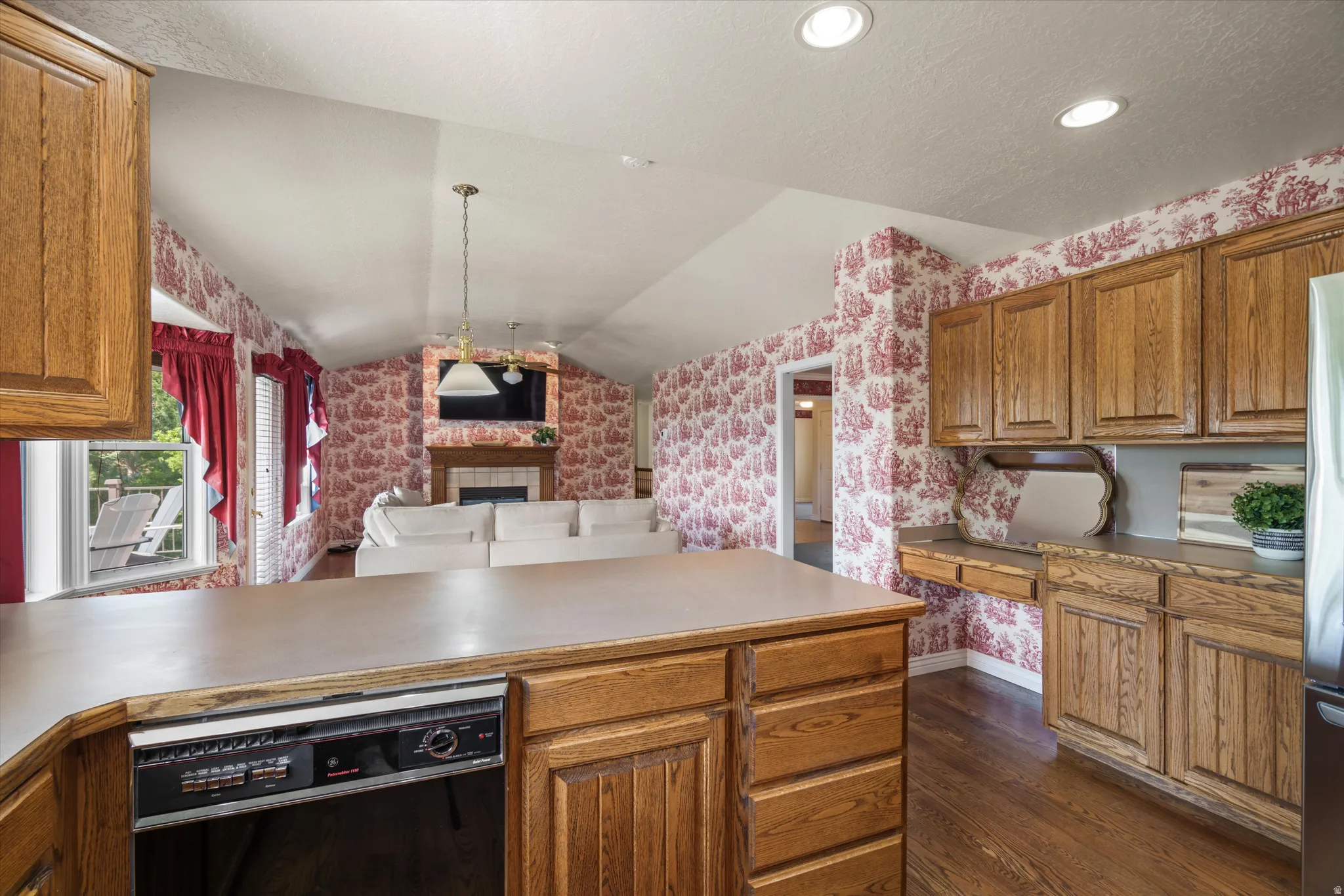 Kitchen island with integrated dishwasher and wood-finish cabinetry, overlooking a living area with a fireplace and vaulted ceilings