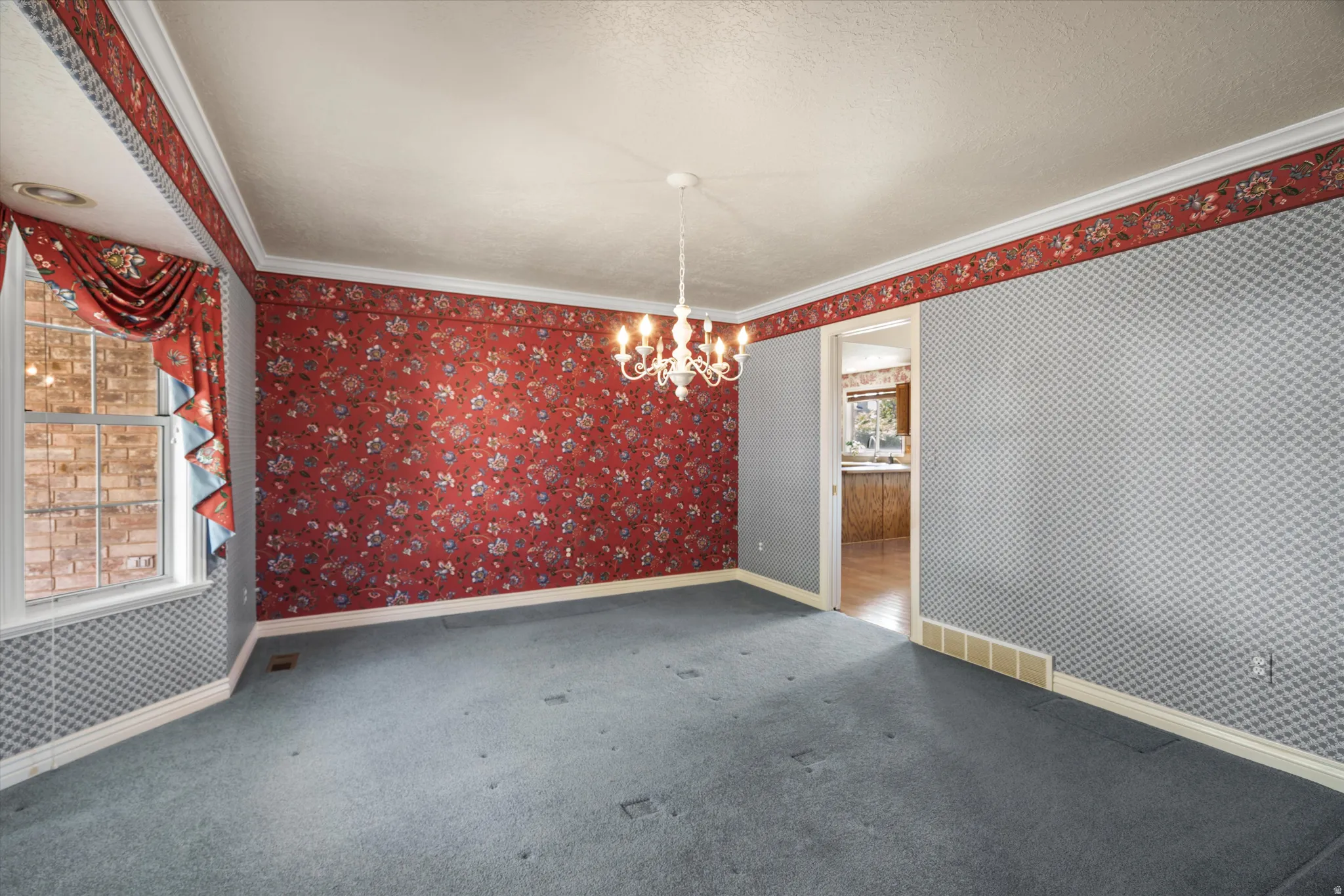 Spacious room featuring patterned wallpaper, crown molding, a chandelier, and a large window with brick exterior visibility