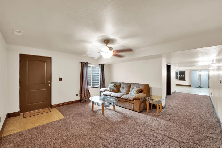 Carpeted living area featuring a ceiling fan with integrated lighting, a window with blinds and drapes, and a brown paneled entry door