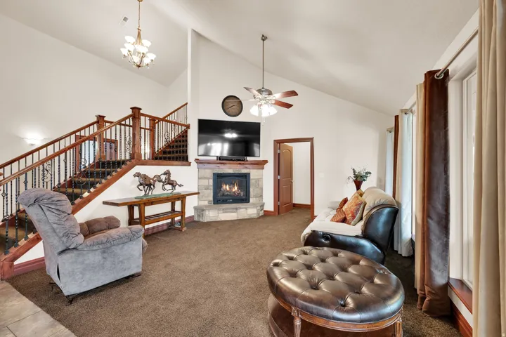 Vaulted ceiling living area featuring a stone-faced fireplace with a wood mantel, a ceiling fan, and a prominent staircase with wood railings and decorative iron balusters