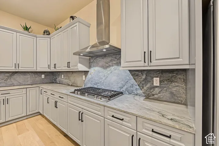 White shaker-style cabinetry, a gas cooktop, stainless steel range hood, stone countertops, and a full-height stone backsplash
