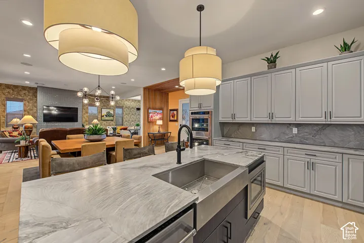 Kitchen island with a large stainless steel farmhouse sink and integrated microwave, light wood-finish flooring, light gray cabinetry with dark hardware, gray stone countertops, and built-in double ovens