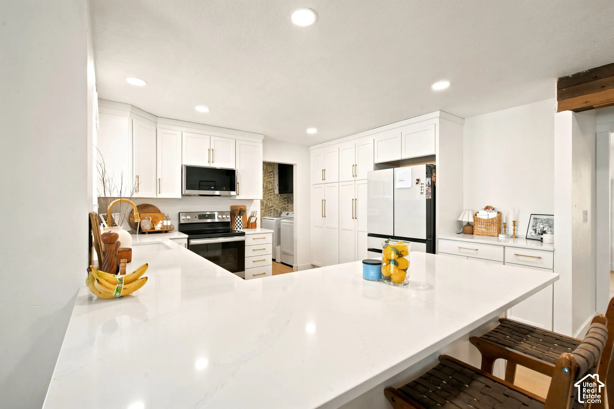 Modern kitchen featuring an expansive white countertop peninsula, recessed lighting, white shaker cabinetry with gold-tone hardware, stainless steel appliances, and a built-in pantry wall