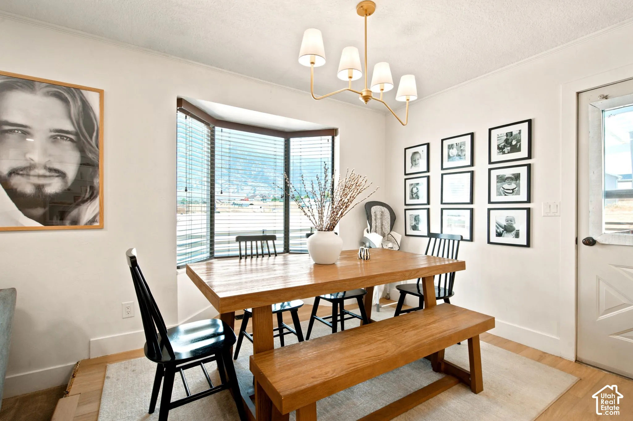 Dining area featuring a prominent bay window with horizontal blinds, light wood-finish flooring, a contemporary gold-finish chandelier with four white shades, and a white paneled door with a window