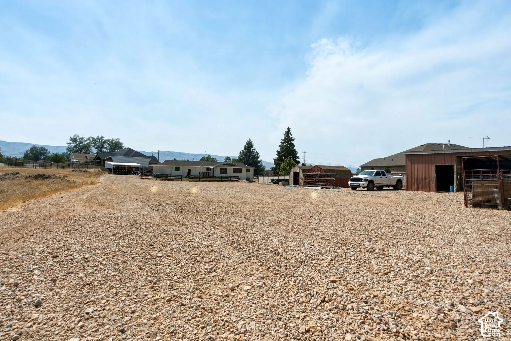 Expansive gravel lot featuring multiple outbuildings, a covered structure, and mature trees on the property perimeter