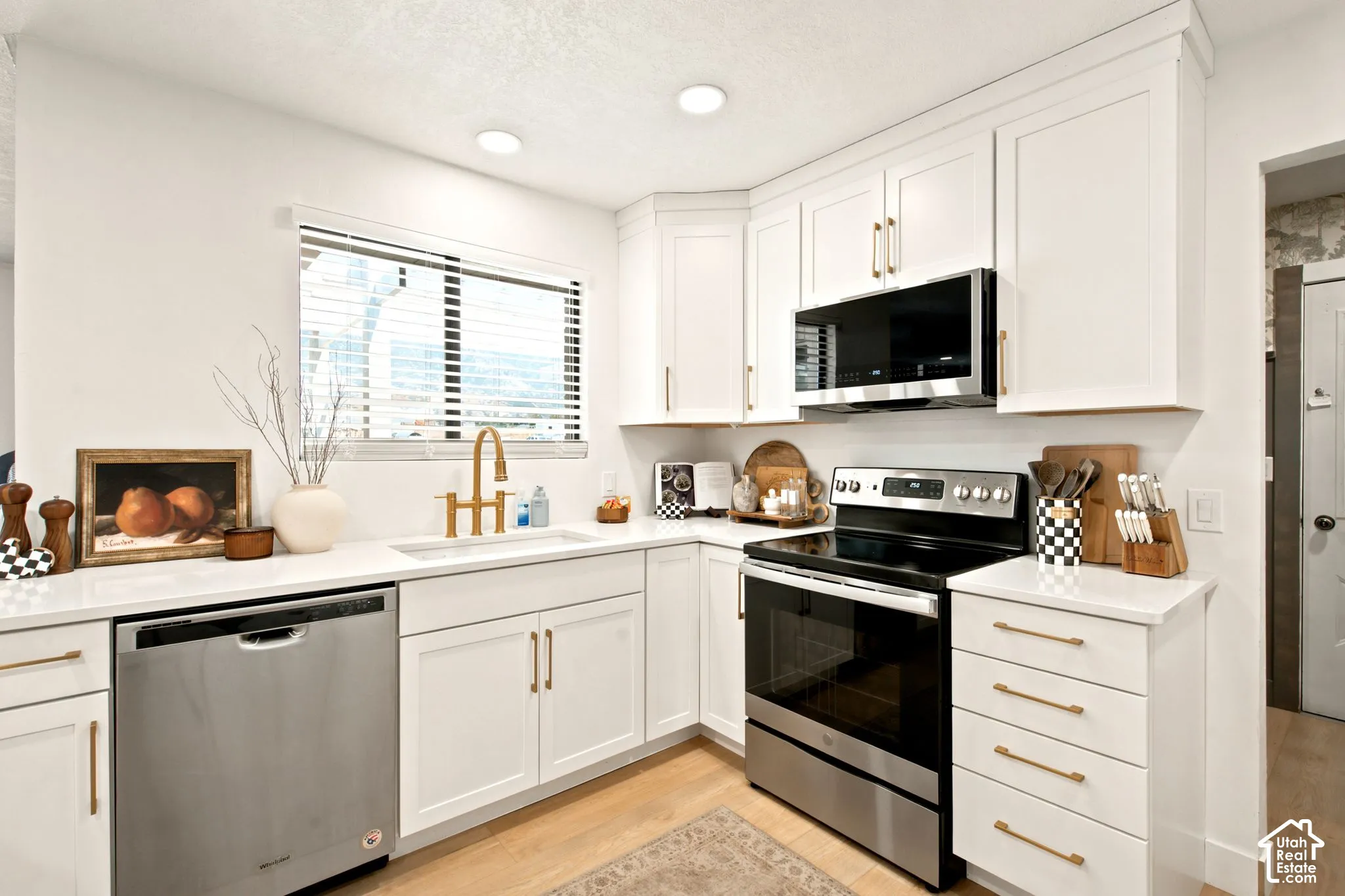 White kitchen featuring recessed lighting, white cabinetry with gold-tone hardware, a stainless steel appliance suite, and light-tone wood-finish flooring