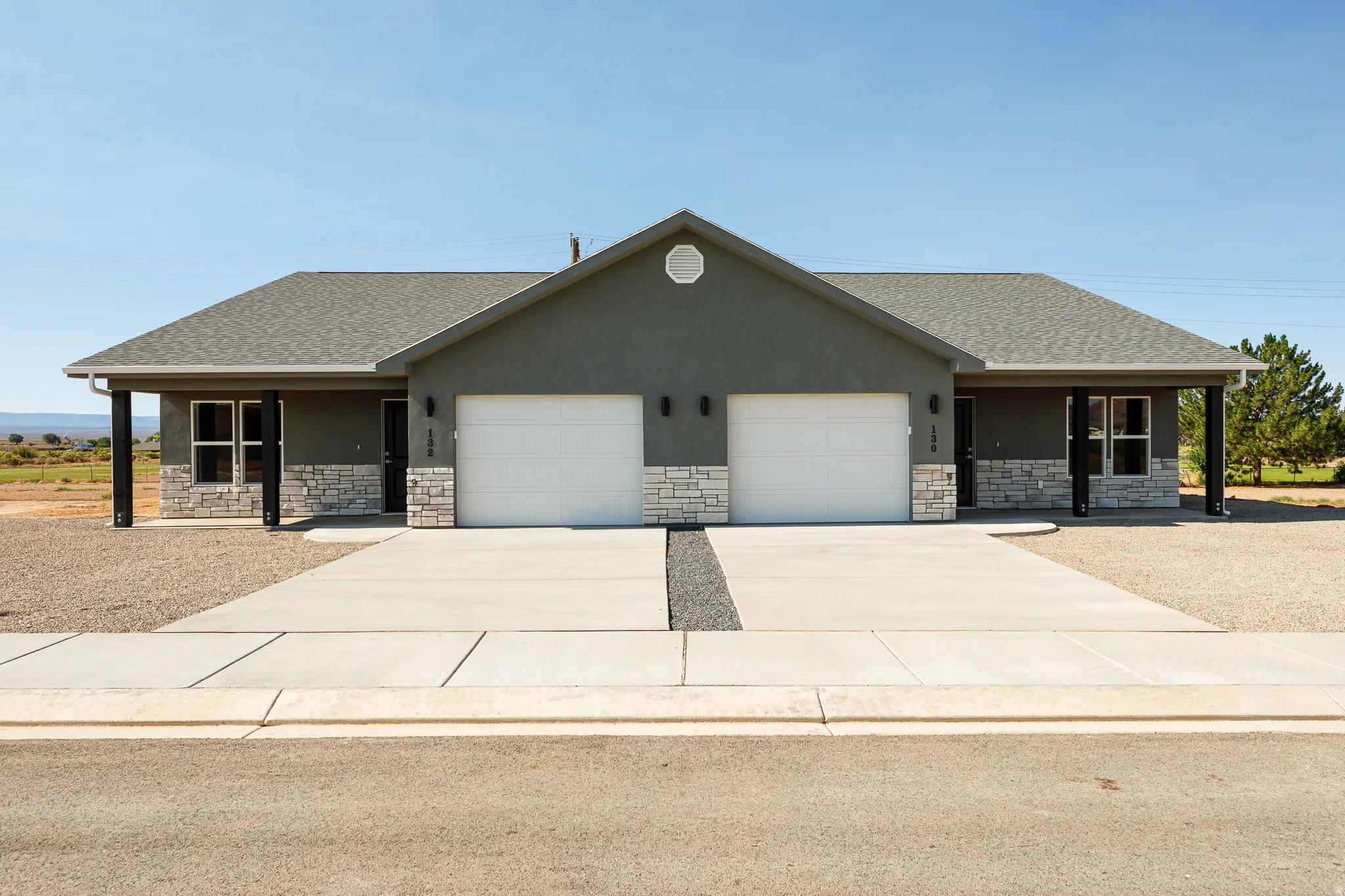 Duplex exterior featuring a gray stucco finish with stone veneer accents, a dark shingle roof, two single-car garages, and covered entryways supported by black columns