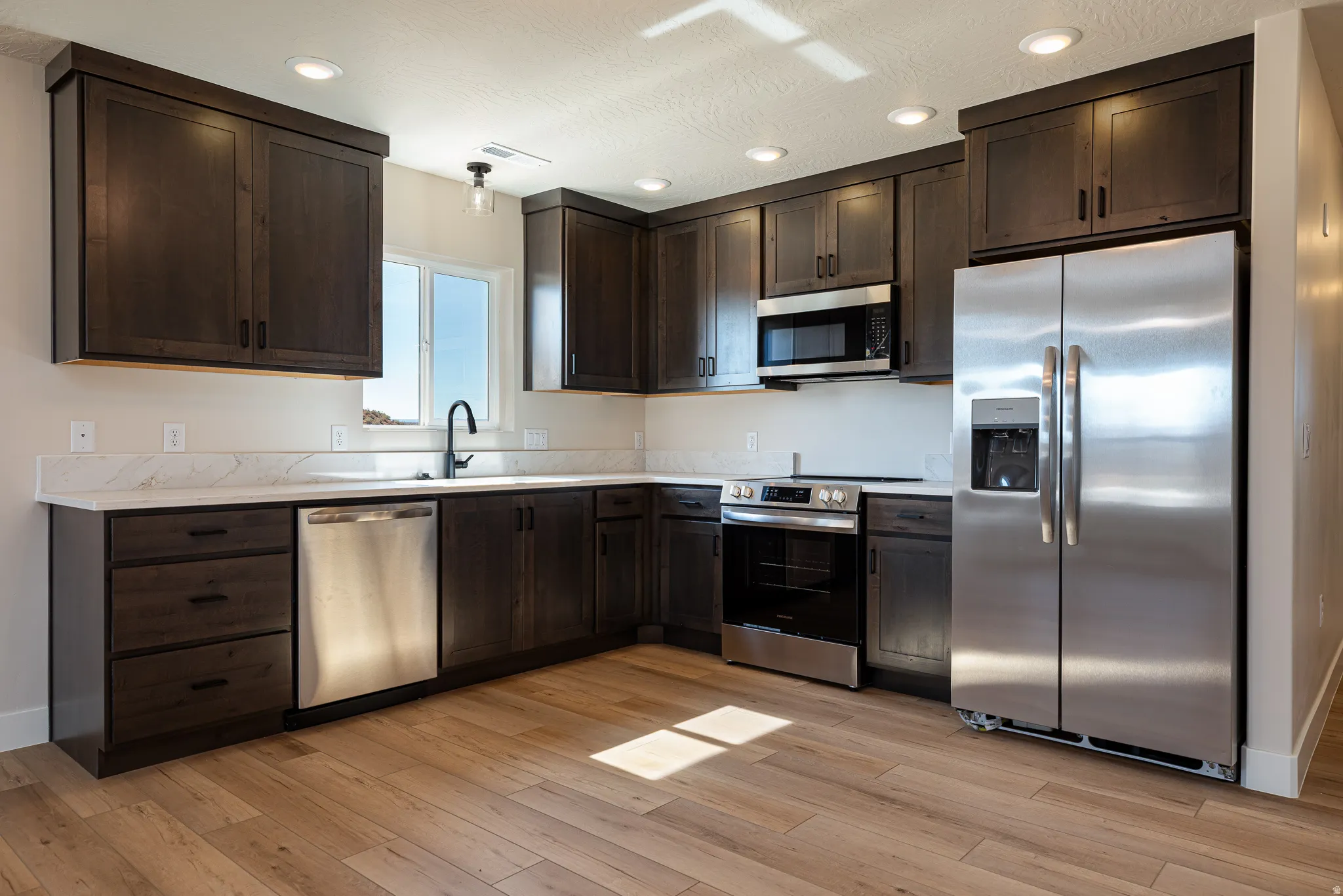 Contemporary kitchen featuring dark wood cabinetry, stainless steel appliances, light-toned countertops, and wood-finish flooring