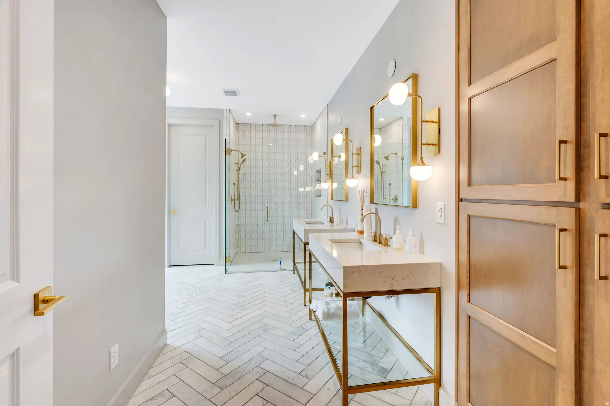 Dual vanity bathroom featuring herringbone tile flooring, a glass-enclosed shower with brass fixtures, and light wood-finish built-in cabinetry