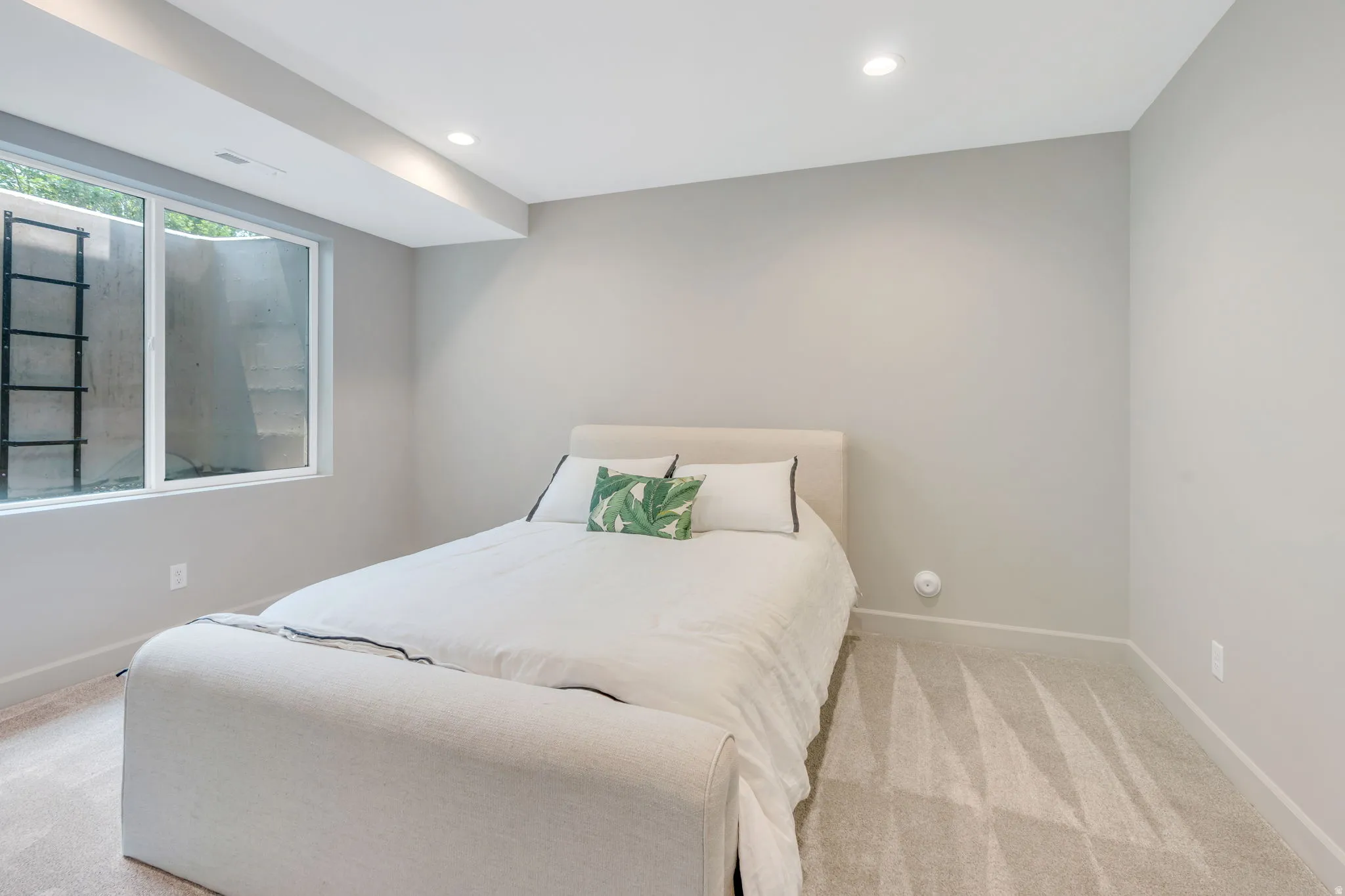 Recessed lighting illuminates this room featuring light gray walls, plush patterned carpeting, and a large egress window