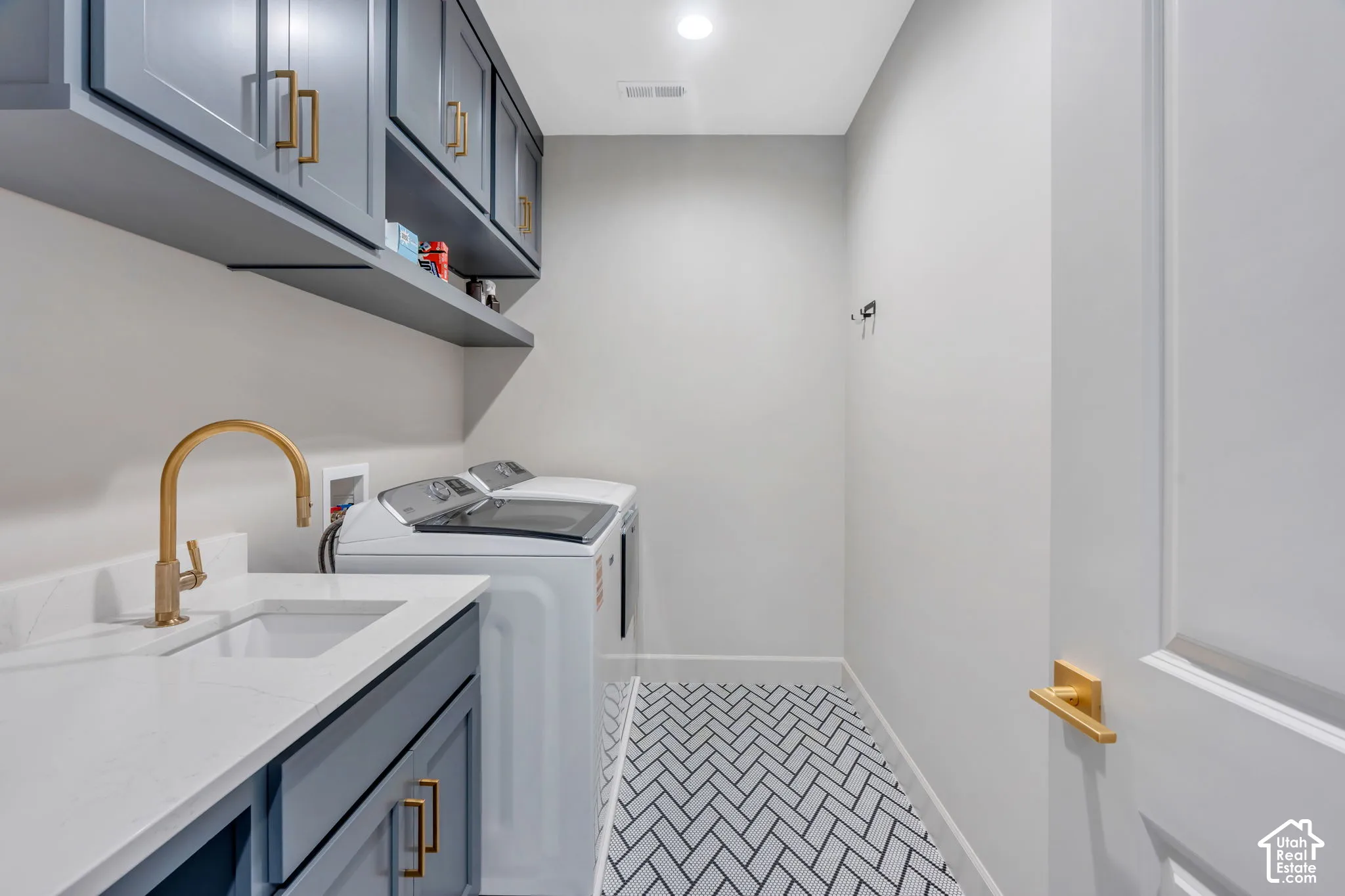 Laundry area featuring a herringbone tile floor, white quartz-finish countertop, undermount sink with a brass-finish faucet, shaker-style cabinetry with brass-finish hardware, and a wall-mounted floating shelf