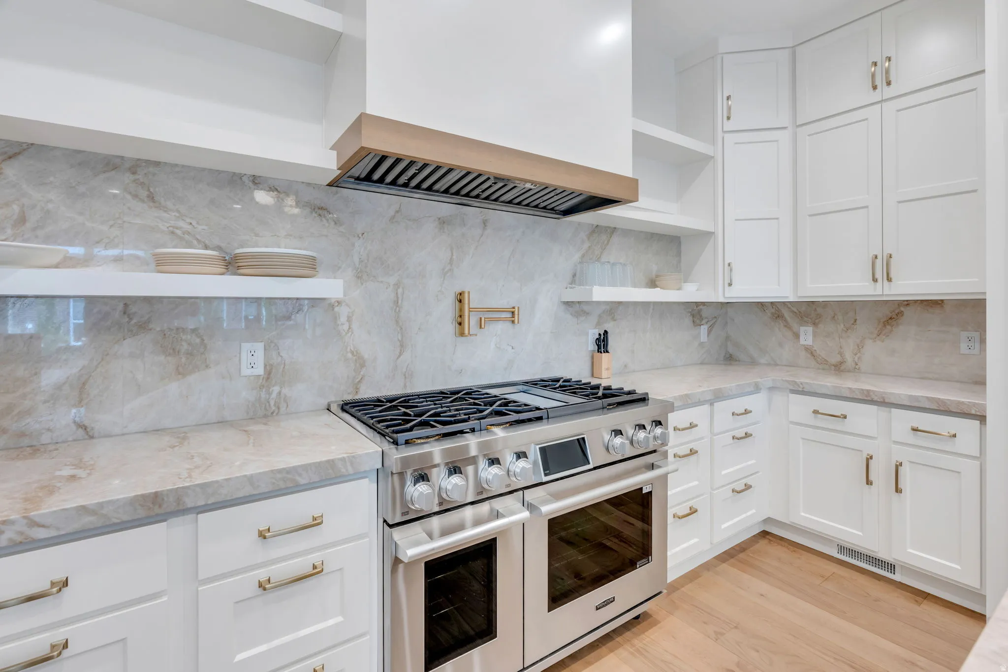 Gourmet kitchen featuring a stainless steel range with a built-in double oven, full-height veined backsplash, matching countertops, white shaker cabinetry with brass hardware, and wood-finish flooring