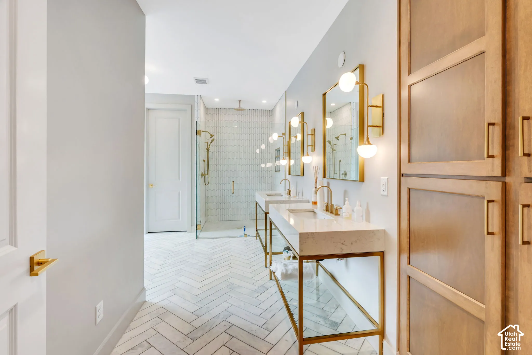 Dual vanity bathroom featuring herringbone tile flooring, a glass-enclosed shower with brass fixtures, and light wood-finish built-in cabinetry