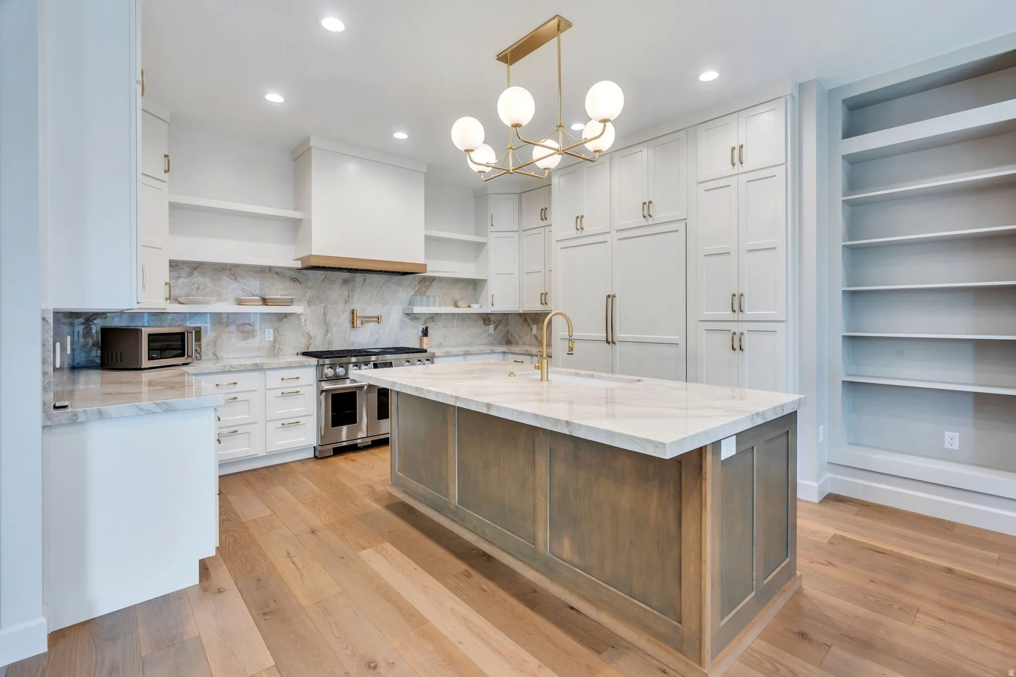 Gourmet kitchen featuring a large central island with a stone countertop, light wood-finish flooring, full-height stone slab backsplash, stainless steel range with custom hood, and extensive built-in cabinetry