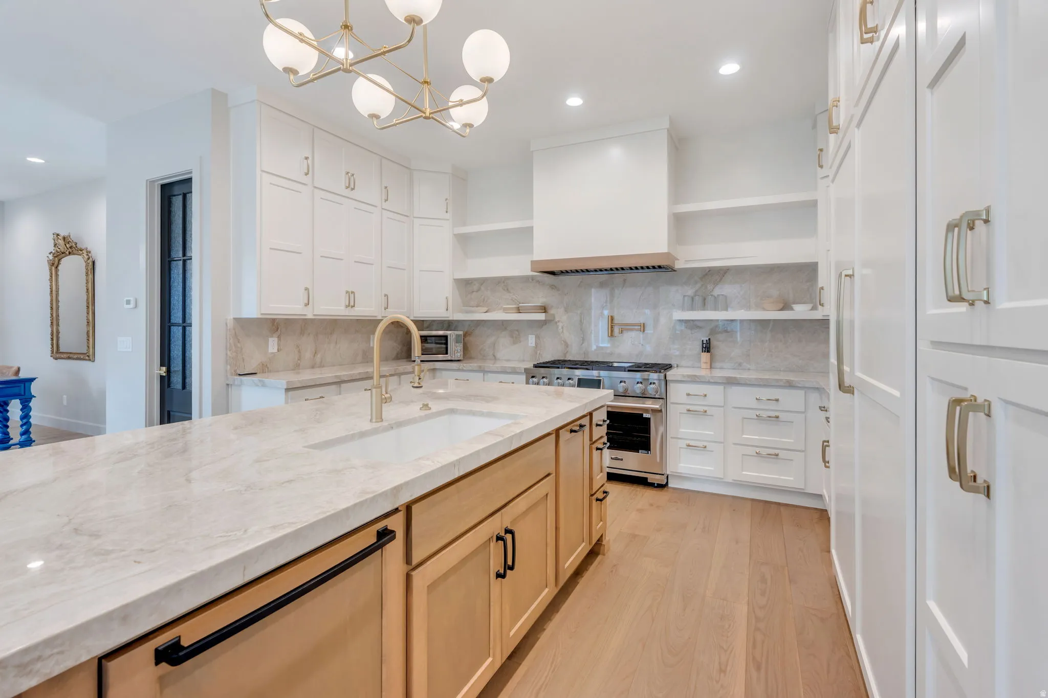 Gourmet kitchen featuring a large central island with integrated sink, stainless steel range with custom hood, extensive white cabinetry, light wood-finish flooring, and a contemporary chandelier