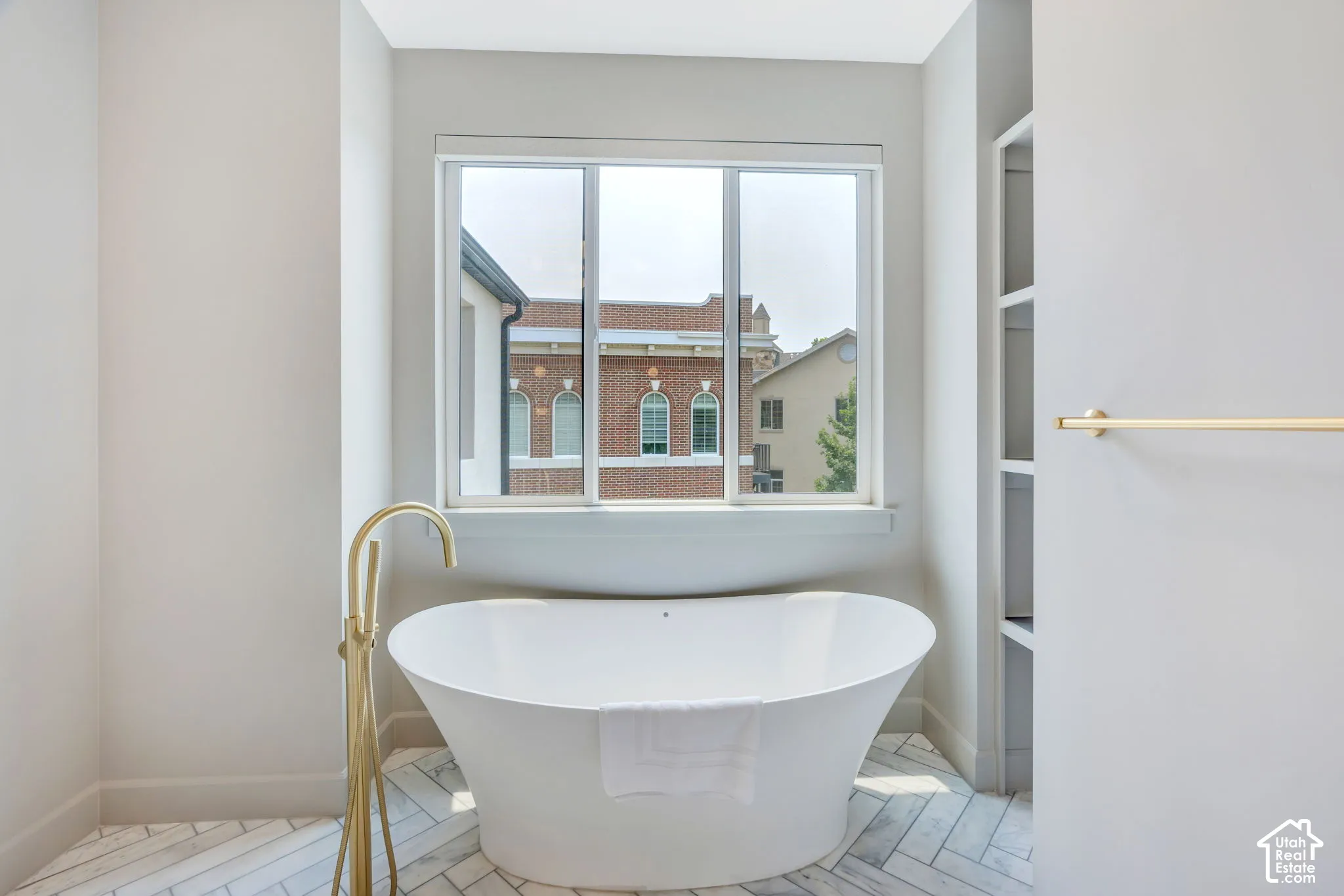Freestanding soaking tub with a floor-mounted gold-tone faucet, herringbone pattern tile flooring, a large window, and built-in shelving