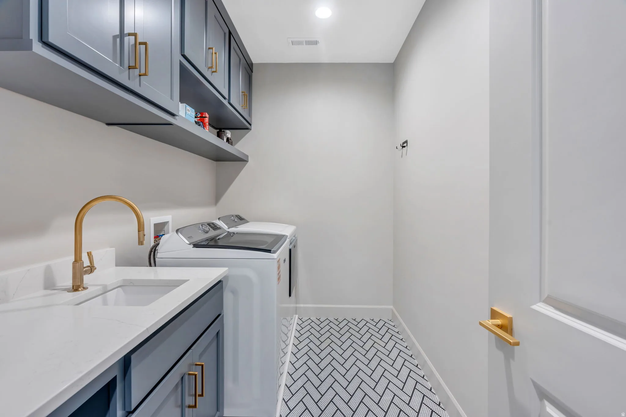 Laundry area featuring a herringbone tile floor, white quartz-finish countertop, undermount sink with a brass-finish faucet, shaker-style cabinetry with brass-finish hardware, and a wall-mounted floating shelf