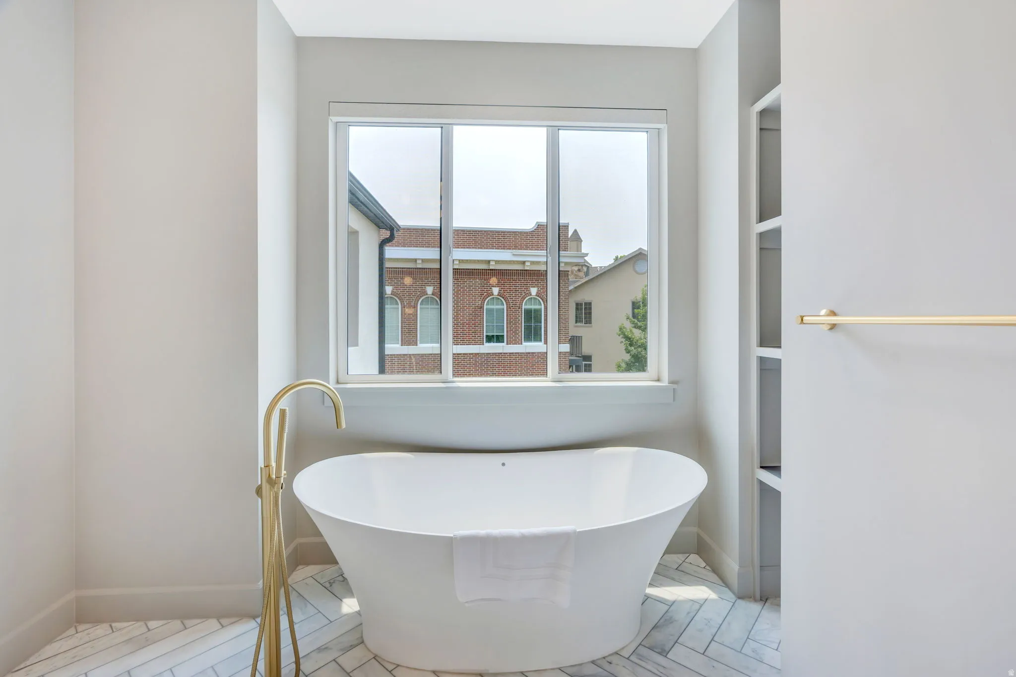 Freestanding soaking tub with a floor-mounted gold-tone faucet, herringbone pattern tile flooring, a large window, and built-in shelving