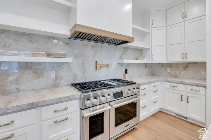 Gourmet kitchen featuring a stainless steel range with a built-in double oven, full-height veined backsplash, matching countertops, white shaker cabinetry with brass hardware, and wood-finish flooring