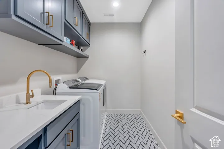 Laundry area featuring a herringbone tile floor, white quartz-finish countertop, undermount sink with a brass-finish faucet, shaker-style cabinetry with brass-finish hardware, and a wall-mounted floating shelf