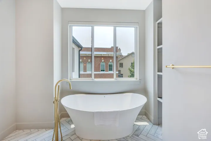 Freestanding soaking tub with a floor-mounted gold-tone faucet, herringbone pattern tile flooring, a large window, and built-in shelving