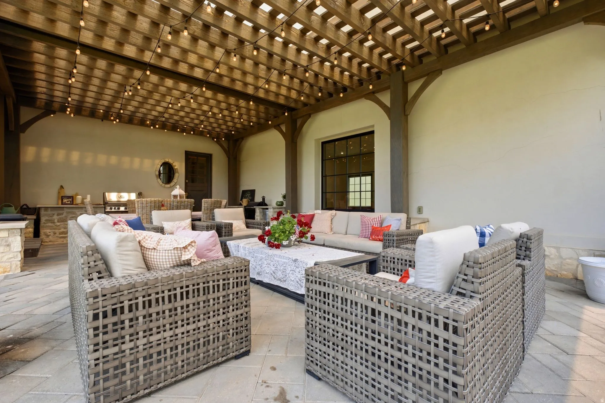 Expansive outdoor patio featuring a wood pergola with string lighting, stone-look paver flooring, a built-in outdoor kitchen with grill, and a dark-framed window