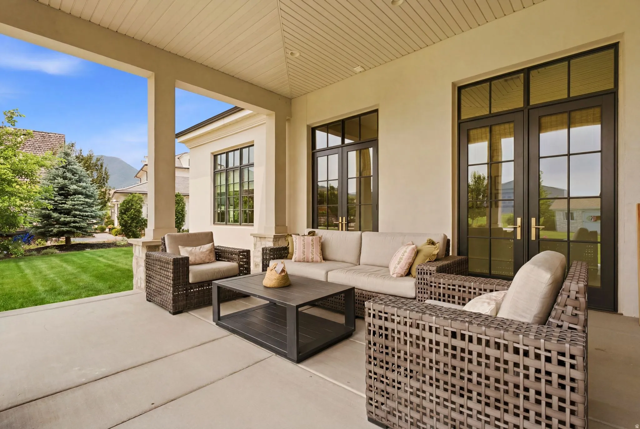 Covered concrete patio with overhead lighting, featuring multiple large grid-patterned glass doors, exterior stucco siding, and a manicured lawn