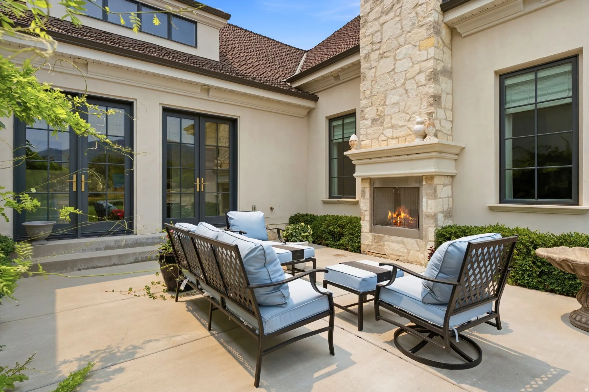 Expansive concrete patio featuring a stone-clad outdoor fireplace, multiple black-framed windows, and two sets of double French doors providing direct access to the interior