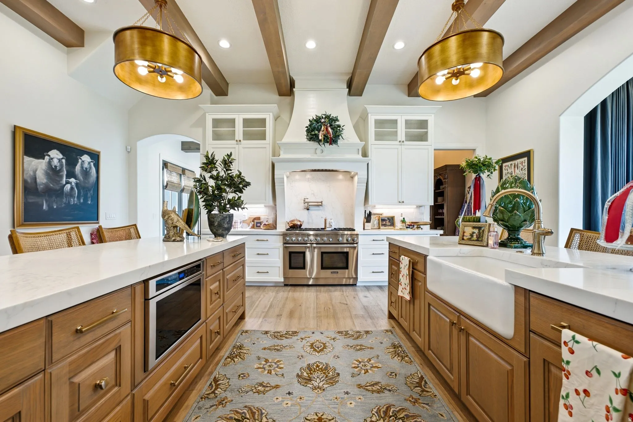 Spacious kitchen featuring two islands with stone countertops, a large farmhouse sink, stainless steel range with custom vent hood, and natural wood ceiling beams
