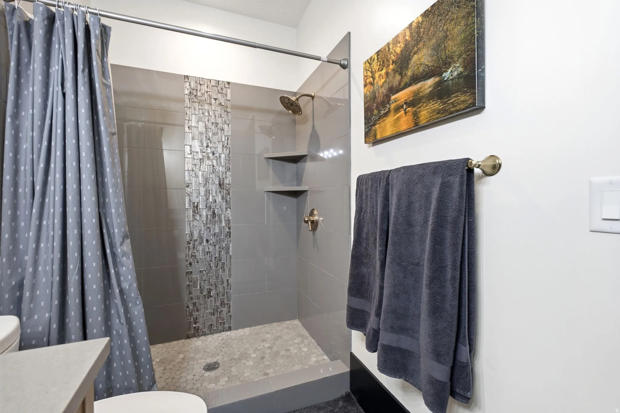 Walk-in shower featuring gray tile walls, a decorative mosaic tile accent strip, and a pebble-style floor