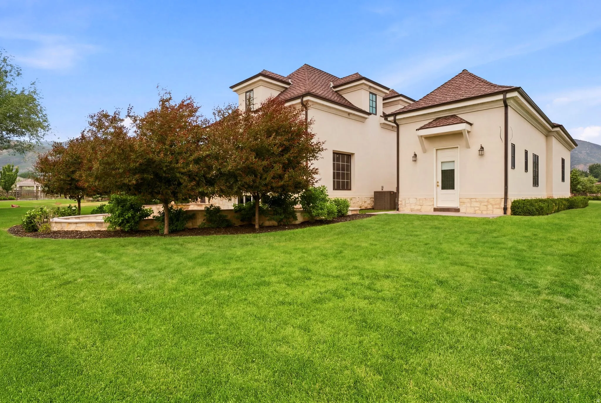 Spacious exterior grounds featuring expansive green lawn, mature trees, stucco facade, stone detailing, and a shingle roof