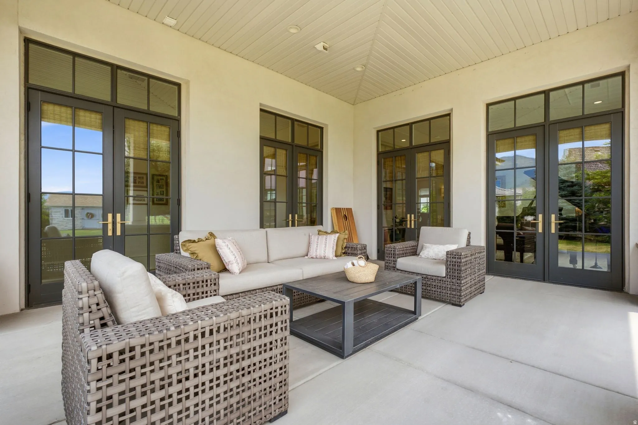 Covered patio featuring a slatted ceiling, light-toned concrete flooring, and multiple sets of black-framed French doors with transom windows and brass hardware