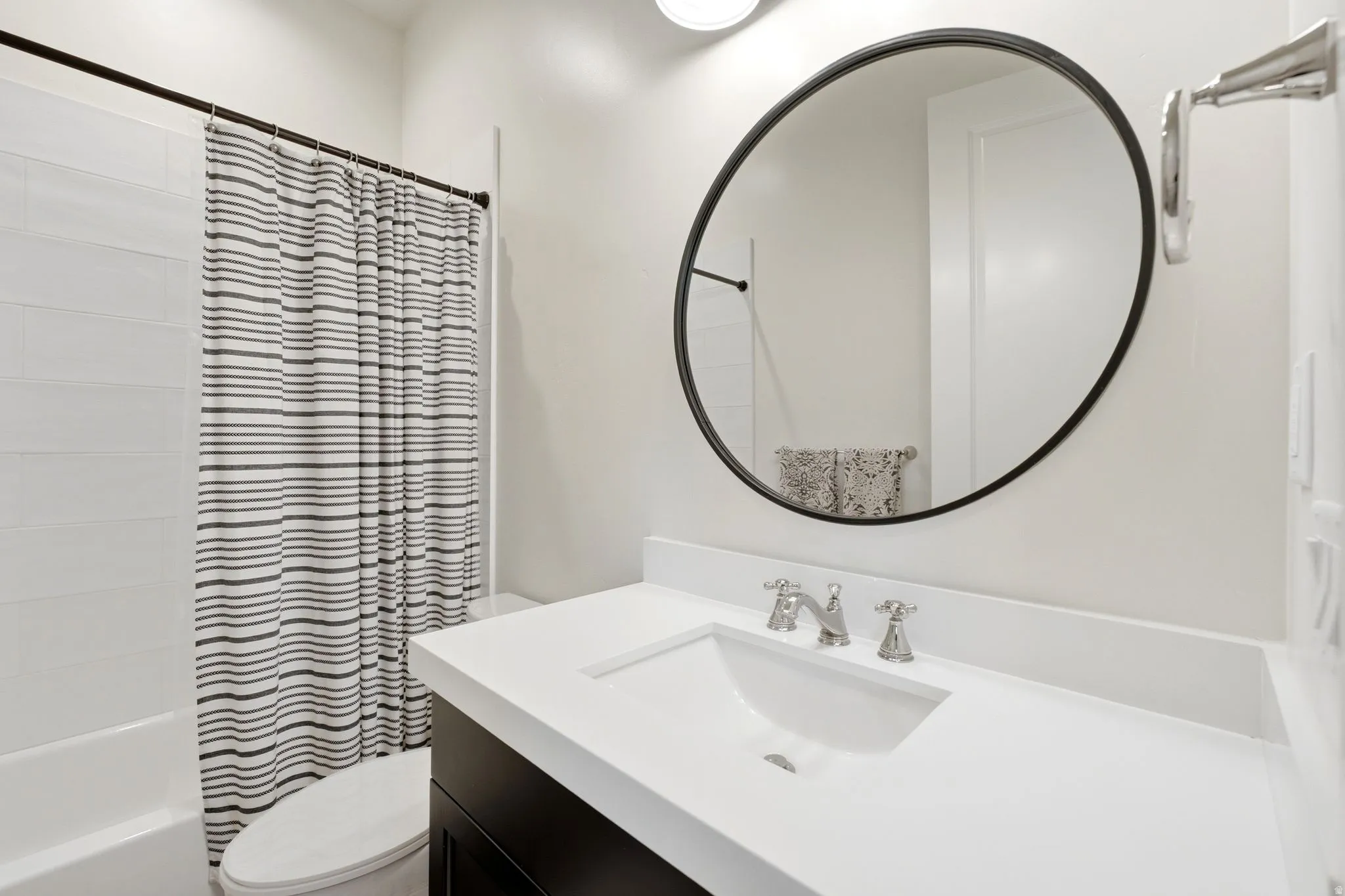 Bathroom featuring a built-in tub with white subway tile surround, a dark wood-finish vanity, white countertop with integrated sink, and a circular black-framed mirror