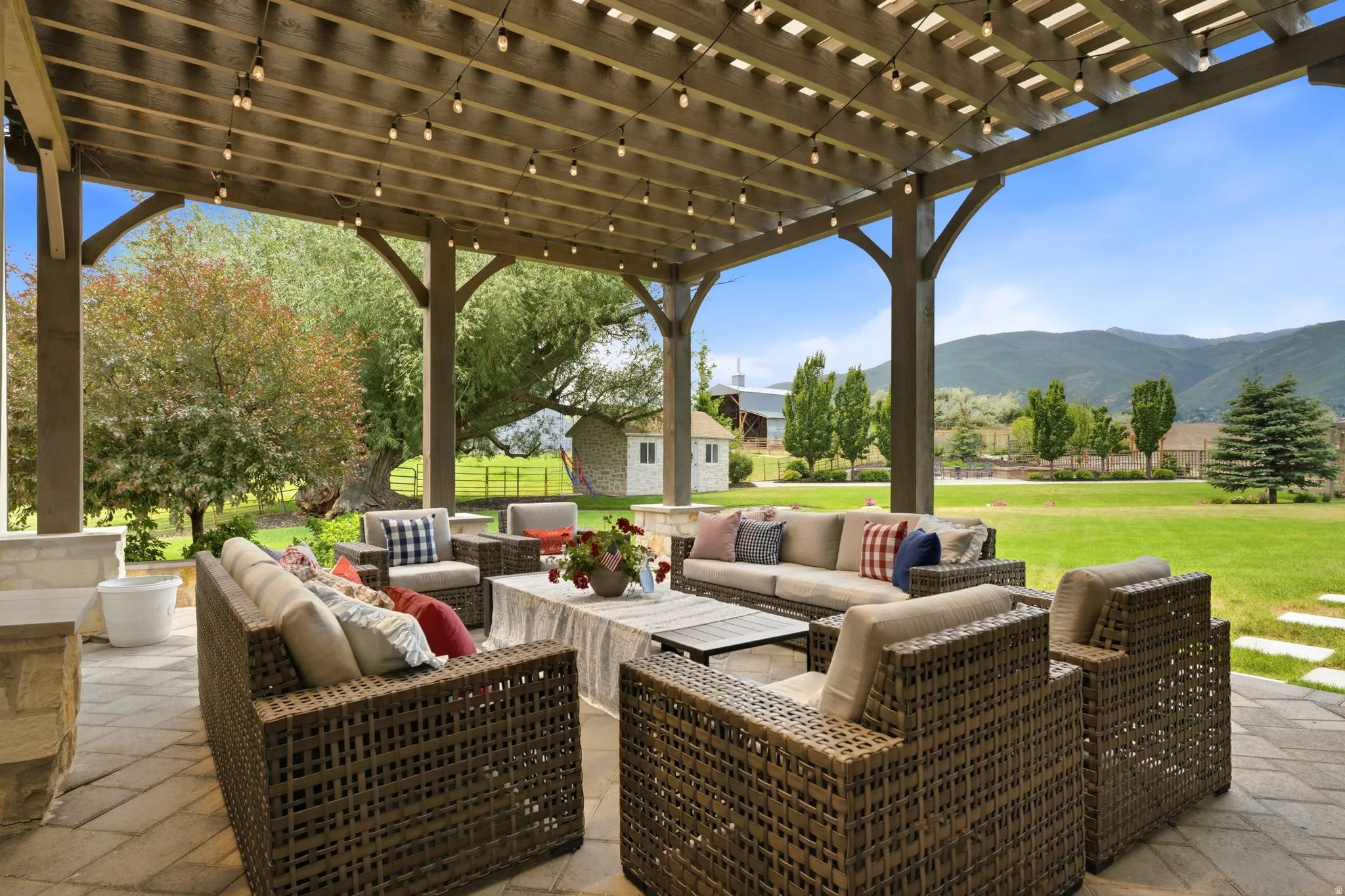 Expansive outdoor patio featuring a wooden pergola with string lights, stone flooring, and a stone-built half-wall