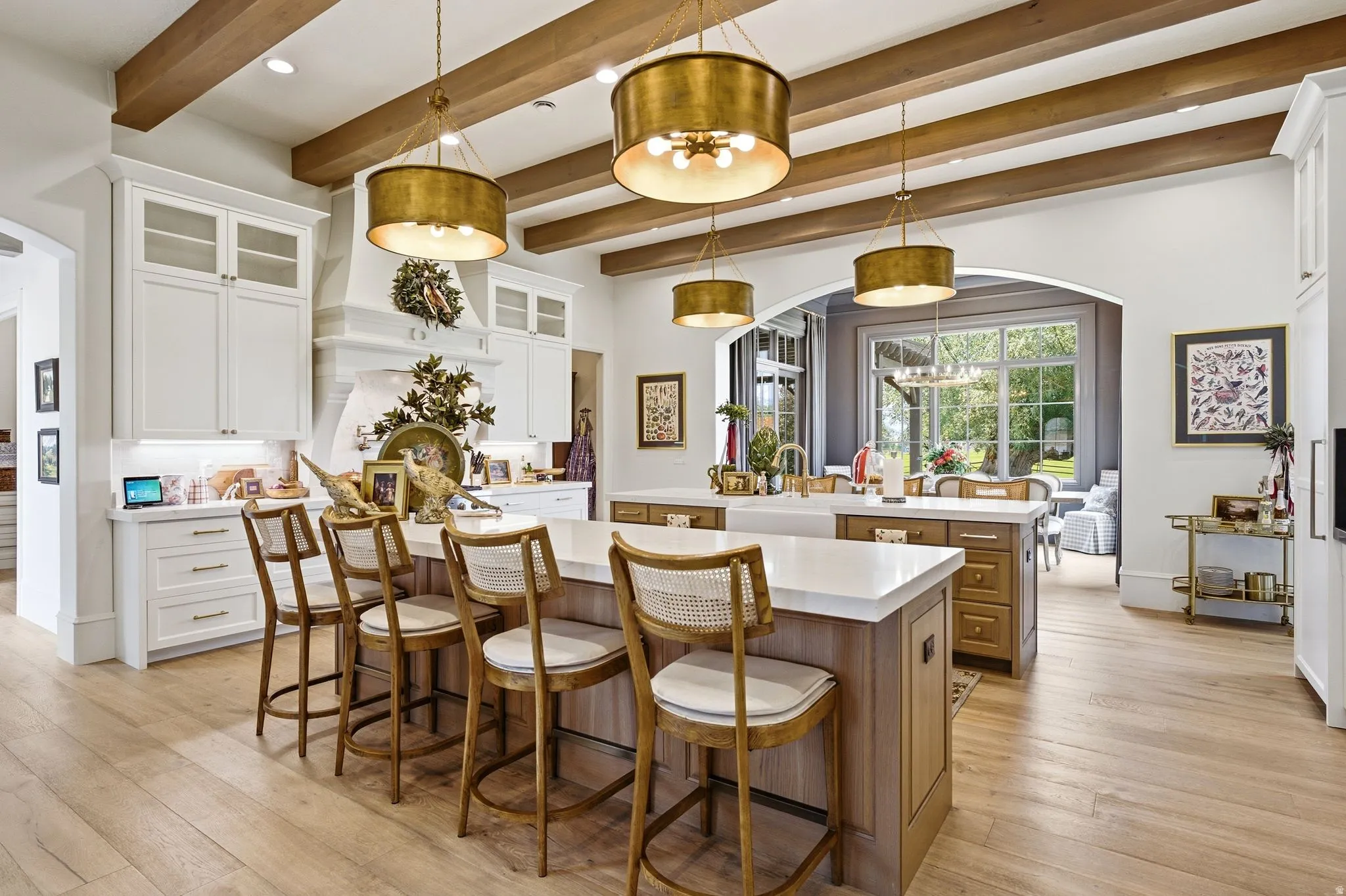 Gourmet kitchen featuring a dual island design, light wood-finish flooring, exposed wood ceiling beams, white cabinetry with glass-front accents, and a built-in range hood